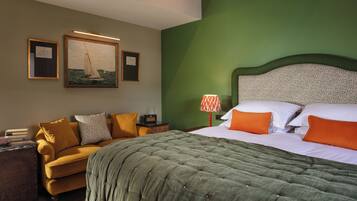 Una habitación de hotel con pared verde, una cama con almohadas blancas y anaranjadas, un edredón acolchado verde, un sillón amarillo, una mesita con un radio y dos cuadros en la pared.
