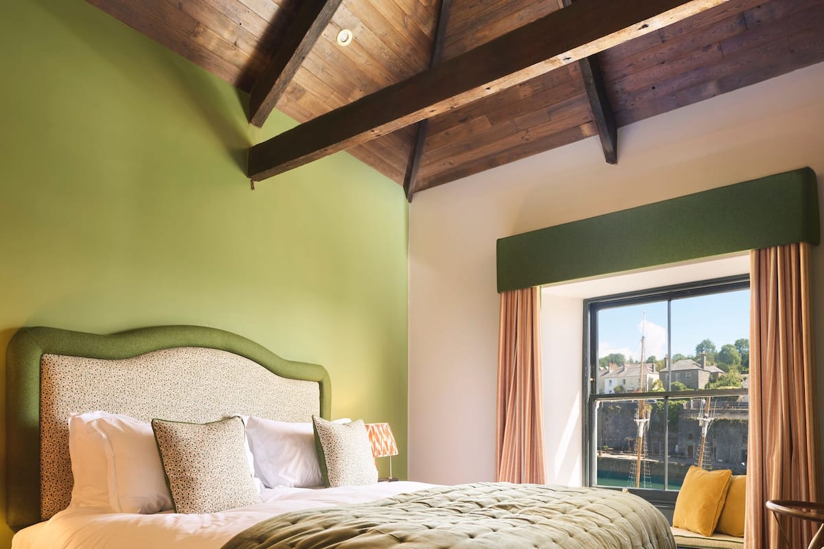 Un dormitorio con techo de madera, una cama grande con edredón verde, una ventana con cortinas y vistas a edificios.