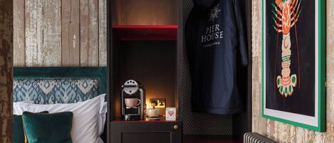 A compact hotel room with a bed, a coffee machine, a hanging robe, and a framed artwork on the wall.