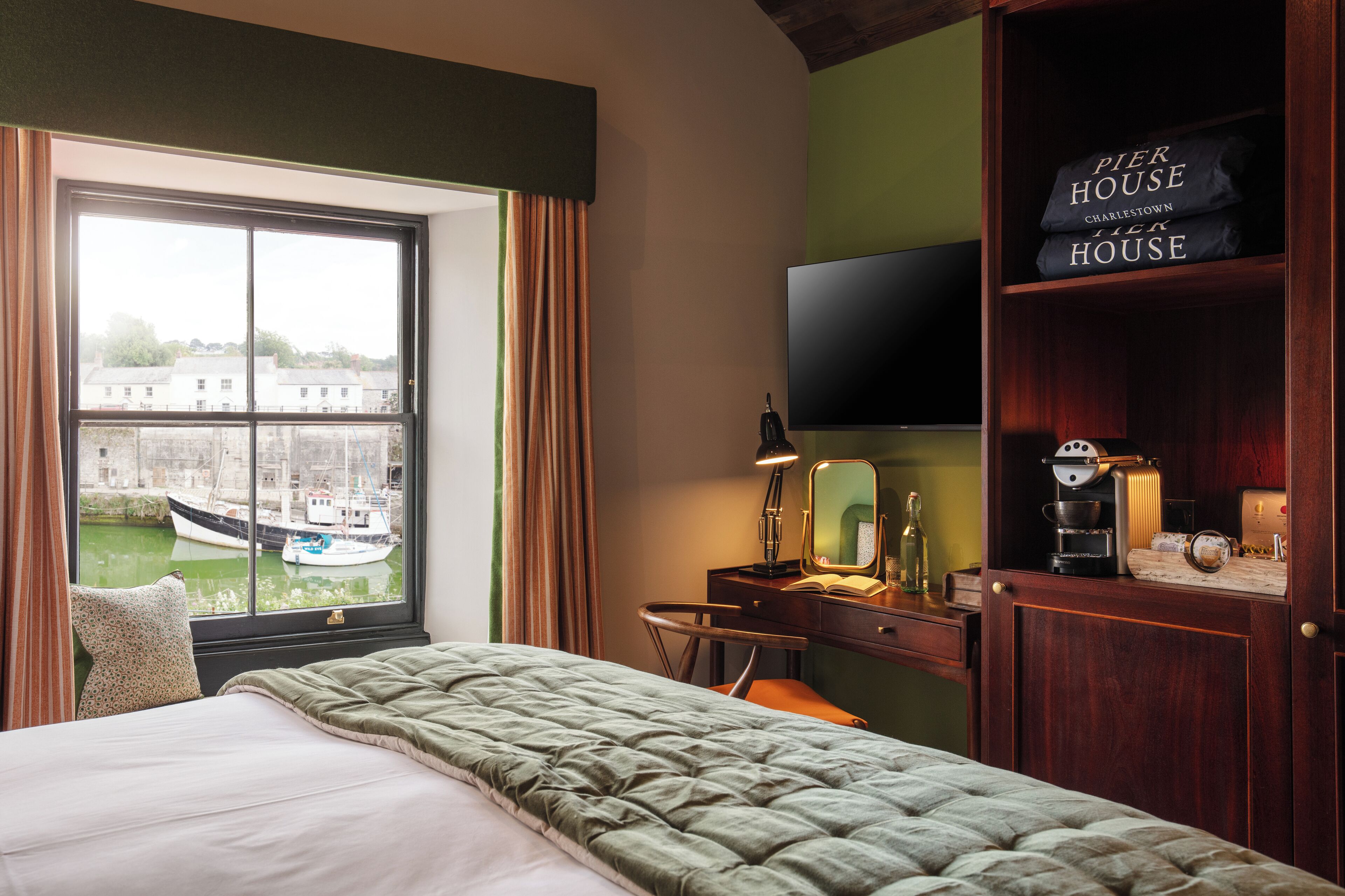 Ein Hotelzimmer mit großem Fenster, einem Bett, einem Schreibtisch mit Lampe, einem Fernseher und einem Regal mit dem Schild "Pier House Charlestown".