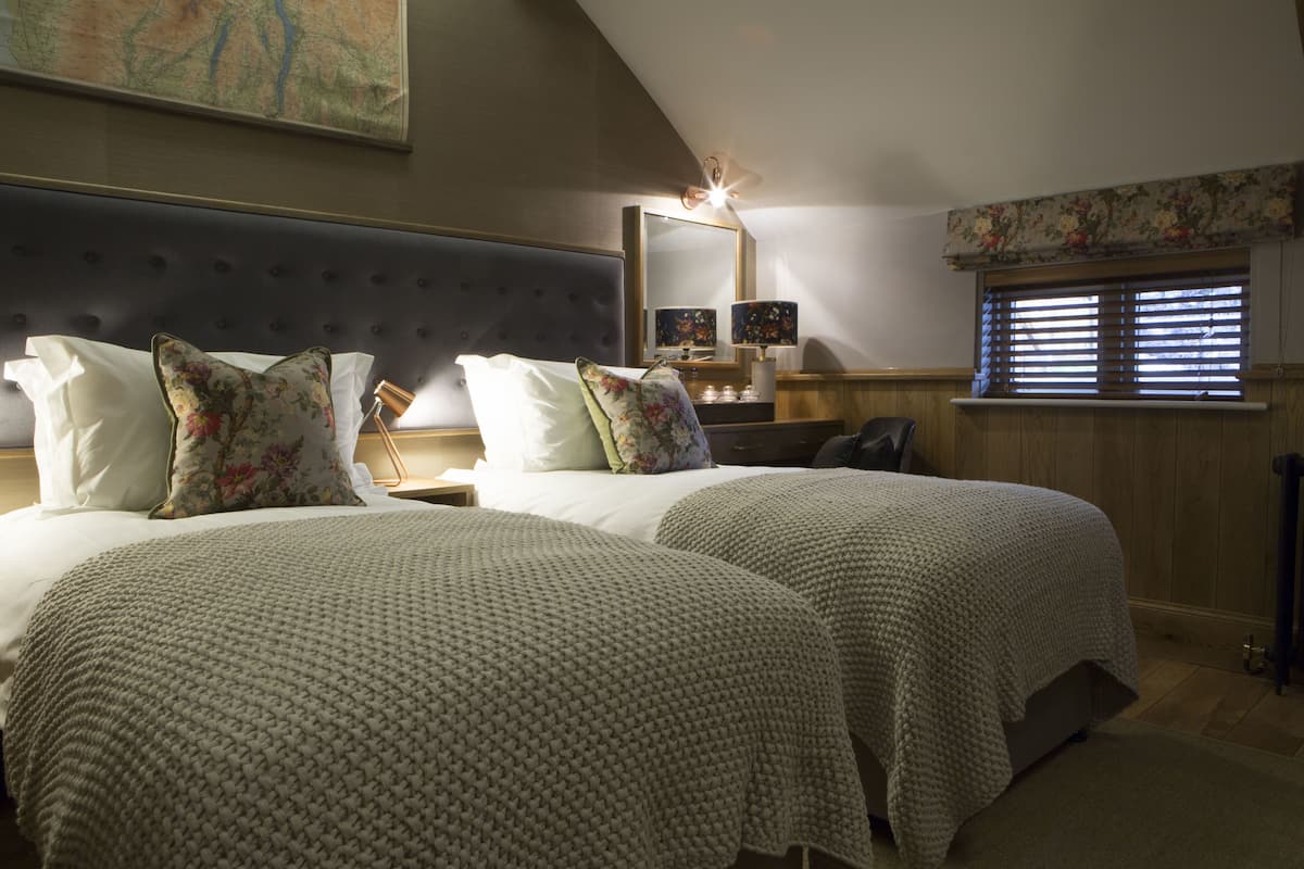 A bedroom with two beds, a large mirror, a nightstand, a lamp, and a window with blinds.