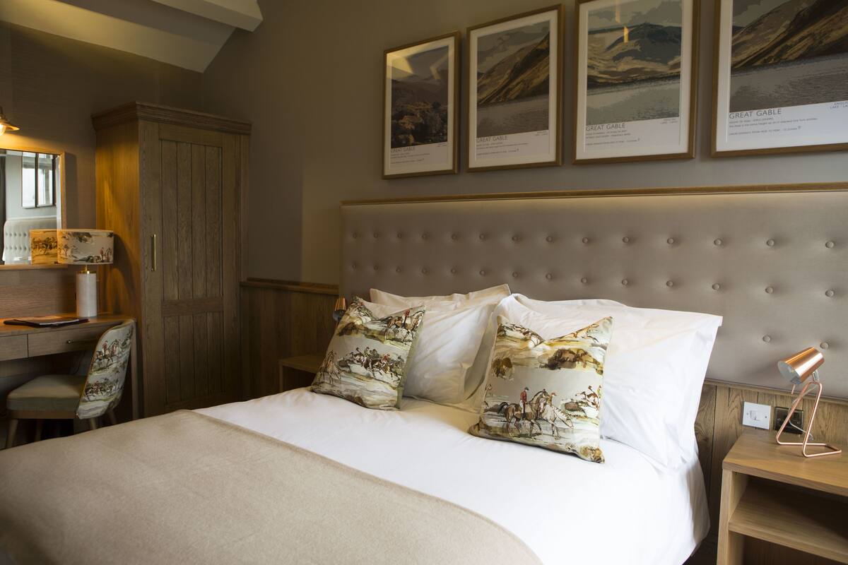 A neatly arranged bedroom with a large bed, two pillows with a landscape print, a bedside table with a lamp, a wooden wardrobe, and framed pictures on the wall.