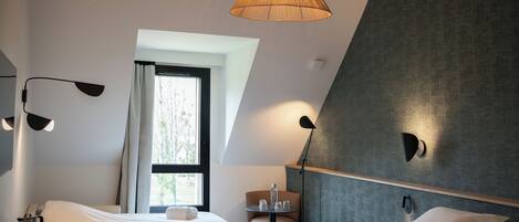 A modern hotel room with two beds, a small table, and a chair. There are two pendant lights and a wall-mounted lamp. A window provides natural light and a view of trees.
