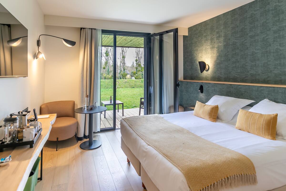 Ein modernes Hotelzimmer mit einem großen Bett, einem Schreibtisch und einem Stuhl. Eine Glastür führt auf einen Balkon mit Blick ins Freie.