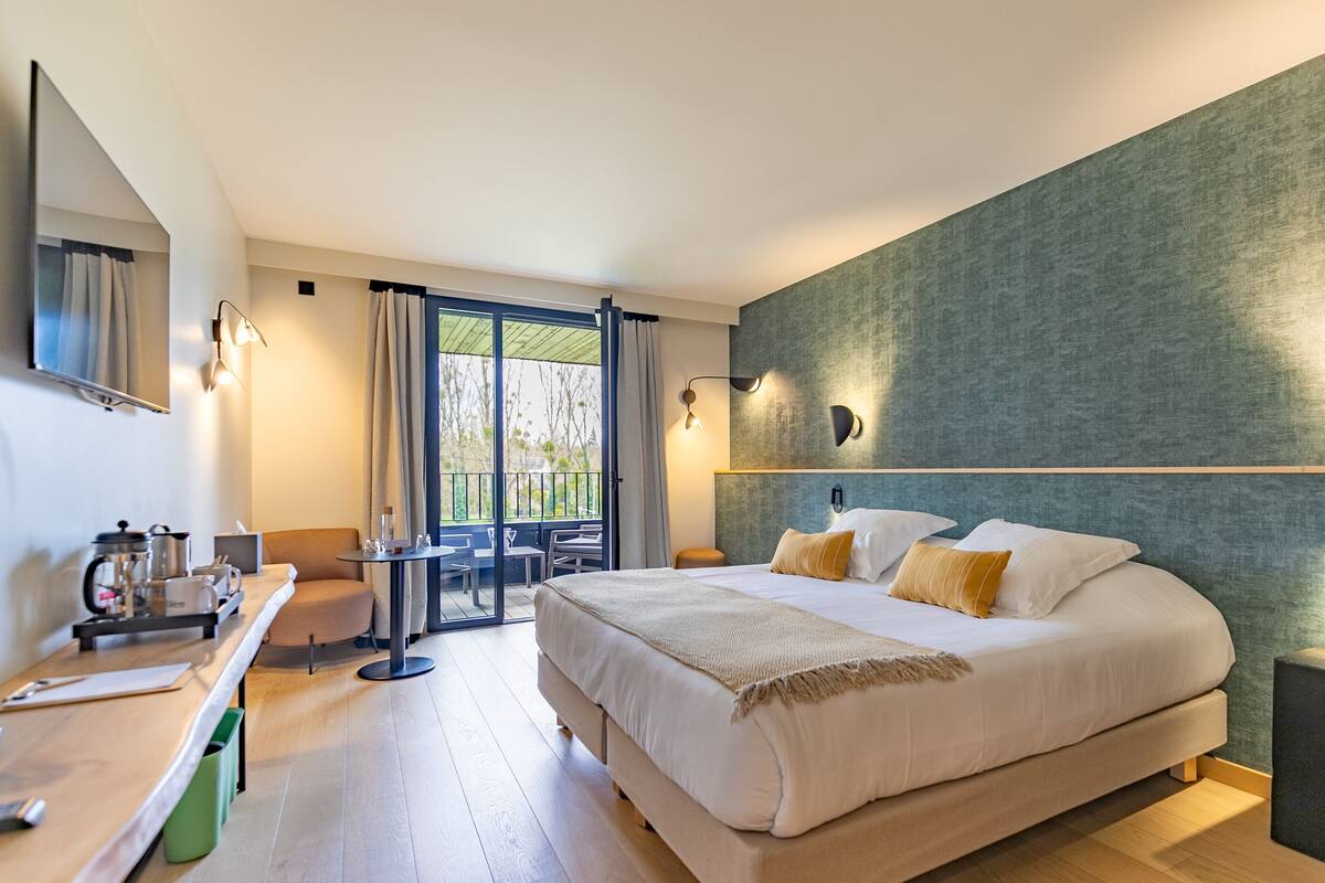 Ein modernes Hotelzimmer mit einem großen Bett, einem Schreibtisch mit Kaffeemaschine, einem Balkon mit Tisch und Stühlen und Blick auf Bäume.