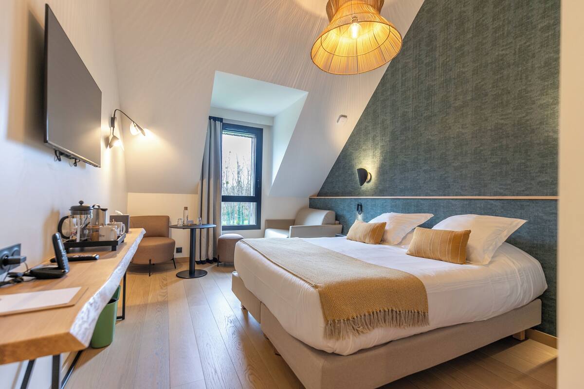 Ein modernes Hotelzimmer mit einem großen Bett, einem Schreibtisch mit Wasserkocher, einem Sessel, einem kleinen Tisch und einem großen Fenster.