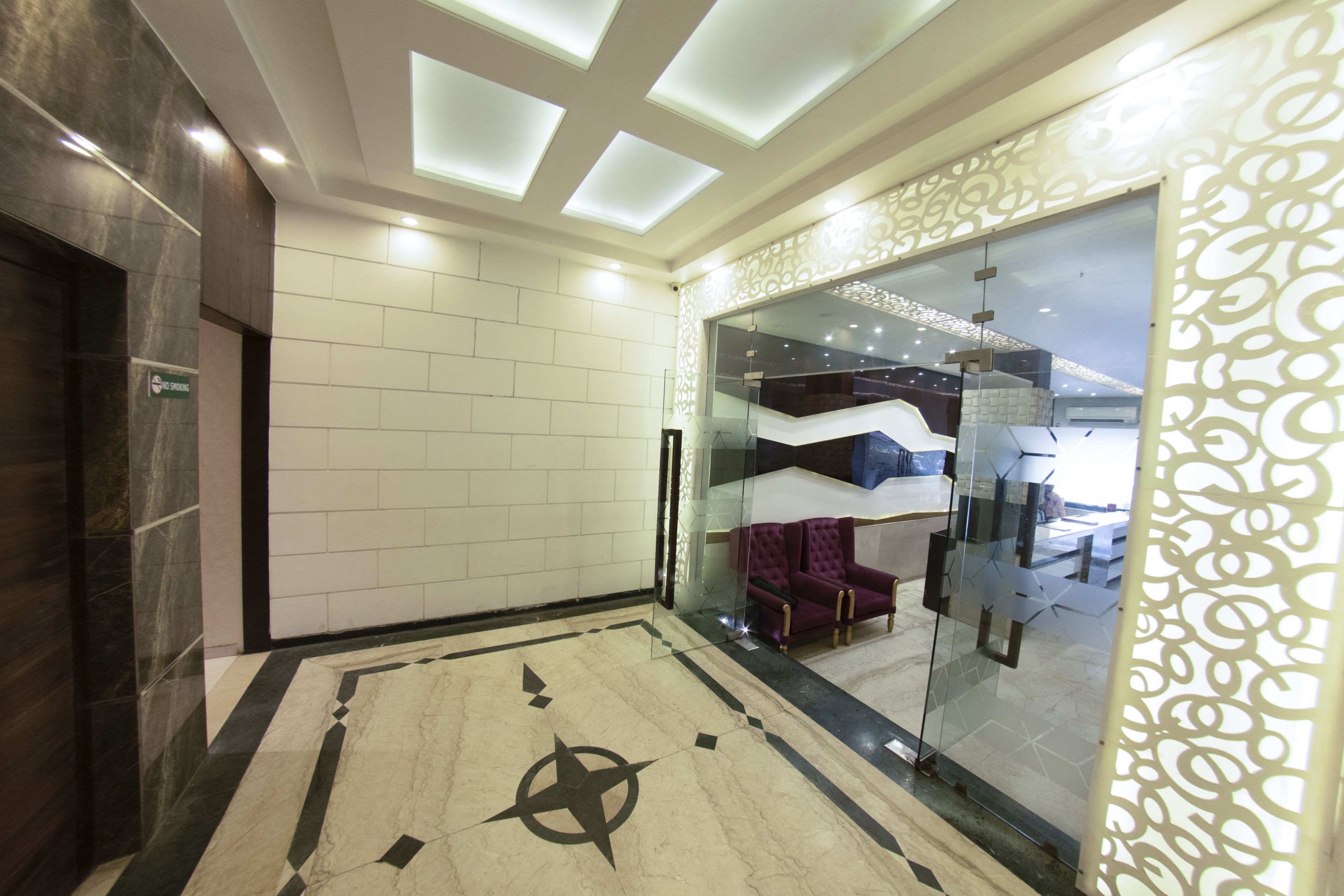 A modern lobby with a geometric floor design, a glass partition with a purple chair, and a ceiling with recessed lighting.