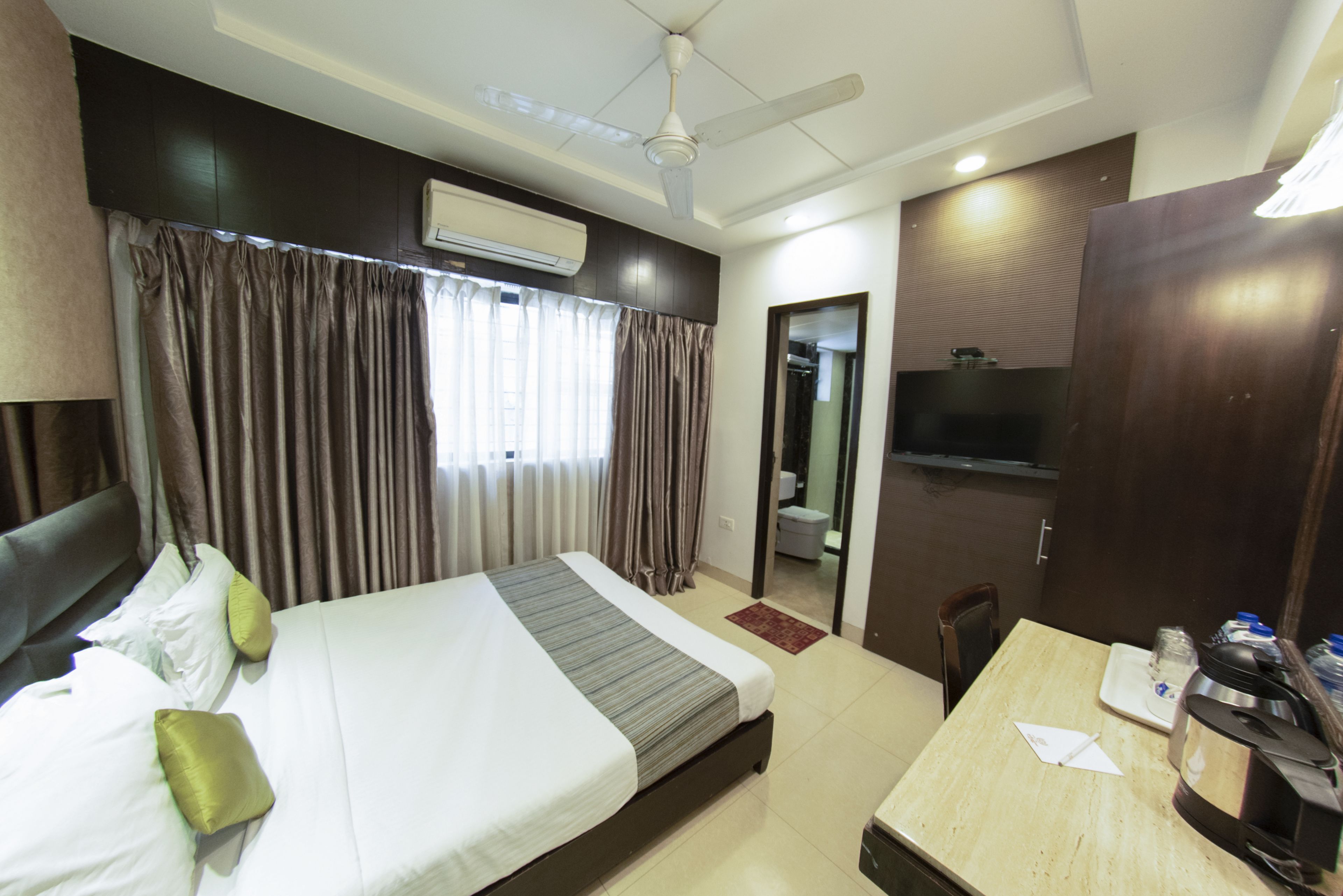 A hotel room with a bed, a desk, a chair, a TV, and an air conditioning unit.