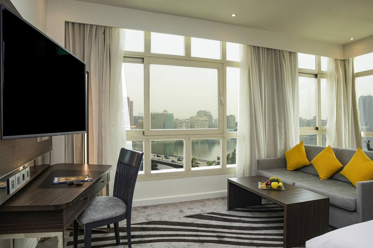 A hotel room with a large window, a flat-screen TV, a dining table, a sofa with yellow pillows, and a coffee table with fruit.