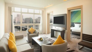 A modern hotel room with a large window offering a city view, a flat-screen TV, a sofa, a coffee table, and a bench.