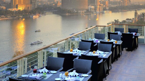A terrace with tables and chairs overlooking a river with boats, with city buildings in the background.
