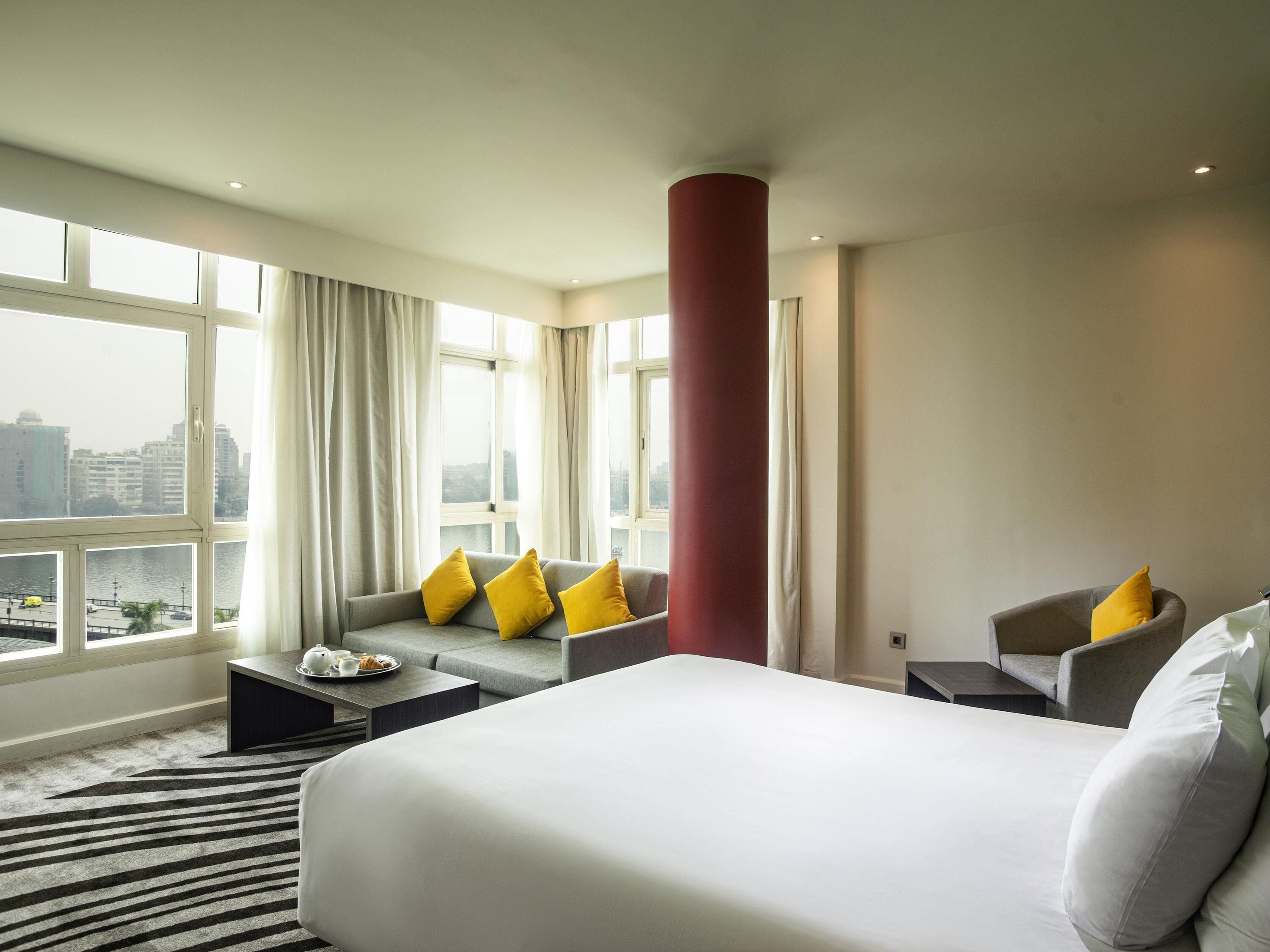 A hotel room with a large bed, a sofa with yellow pillows, a red pillar, and a view of the city through large windows.