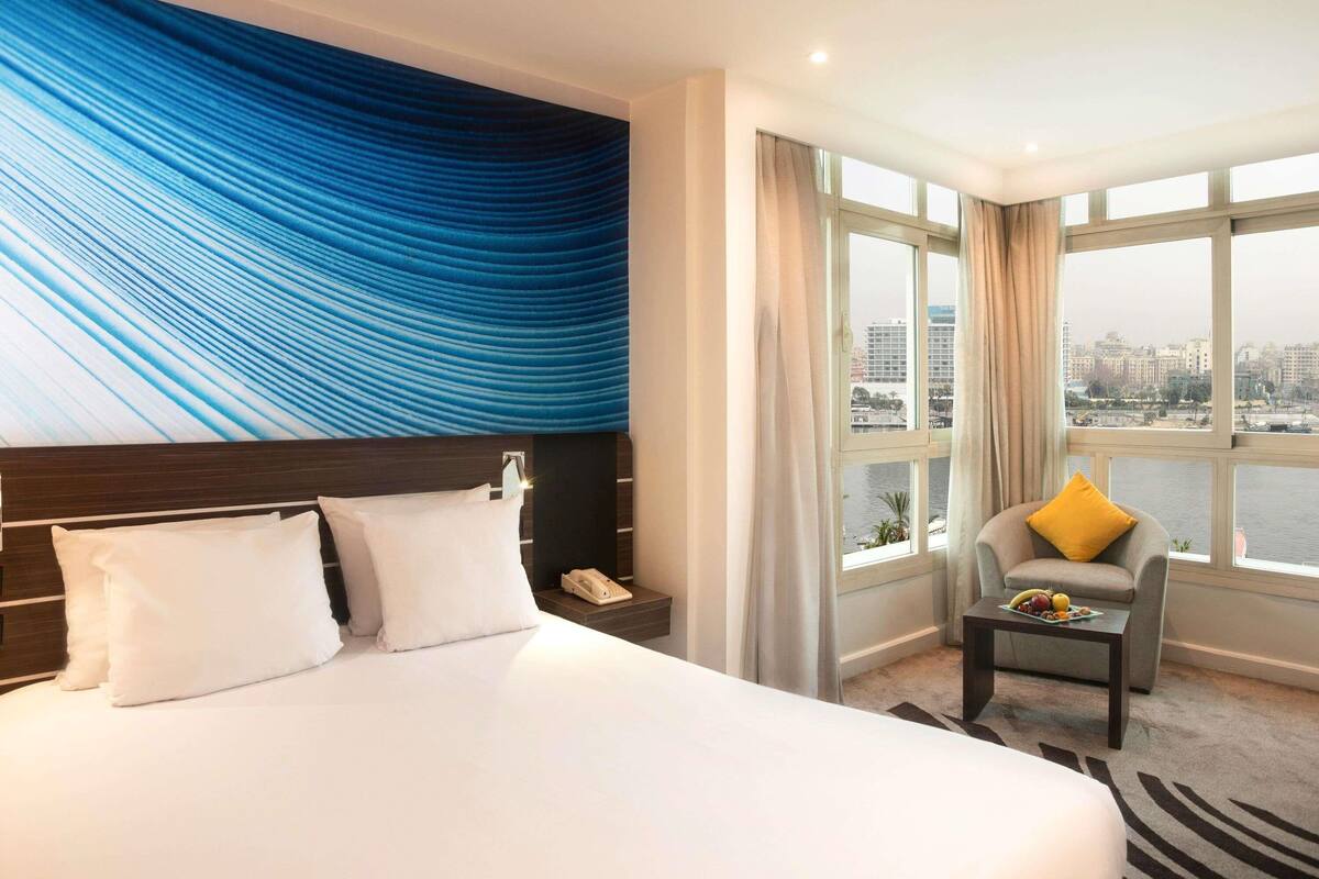 A hotel room with a large bed, a desk with a chair, a view of the city through large windows, and a blue textured wall.
