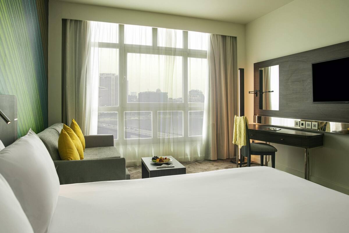 A hotel room with a bed, a sofa, a desk, a chair, a TV, and a view of the city through large windows.