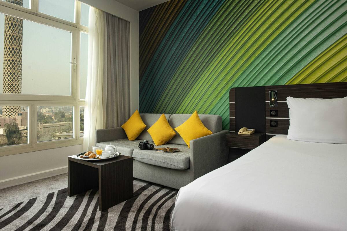 A hotel room with a bed, a sofa with yellow pillows, a small table with breakfast items, and a view of a cityscape through a window.
