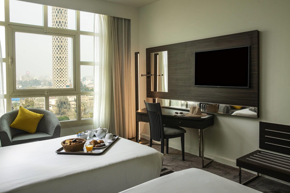 Habitación de hotel con cama, escritorio, silla, televisor y ventana con vista a la ciudad.