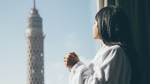 A person in a white robe standing by a window with a cityscape view, including a prominent tower.