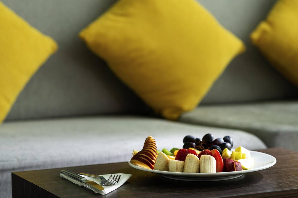 A plate of fruit on a table with a fork and napkin, in front of a couch with yellow pillows.