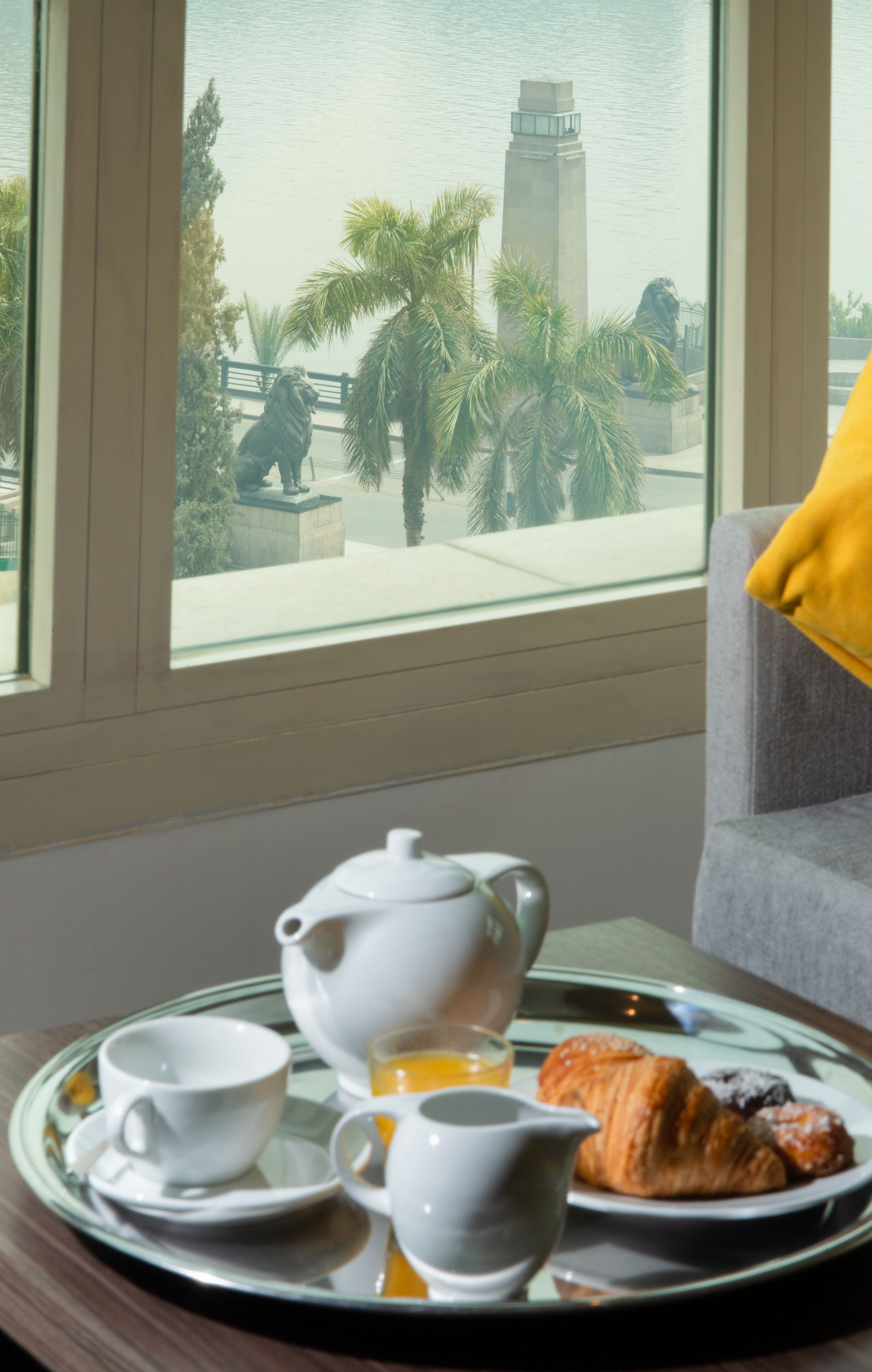 Una bandeja de desayuno con tetera, tazas y pasteles sobre una mesa junto a una ventana con vista a una estatua y palmeras.