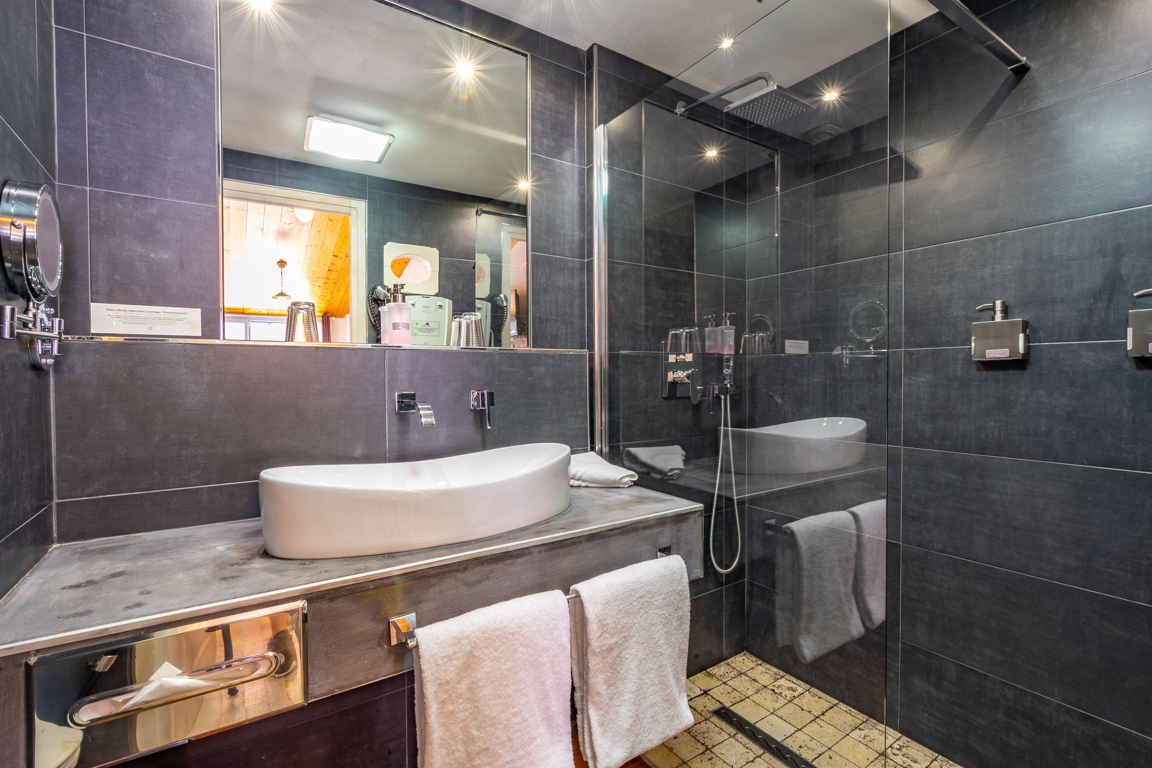 A modern bathroom with a large mirror, a white sink, a glass shower enclosure, and a tiled floor.