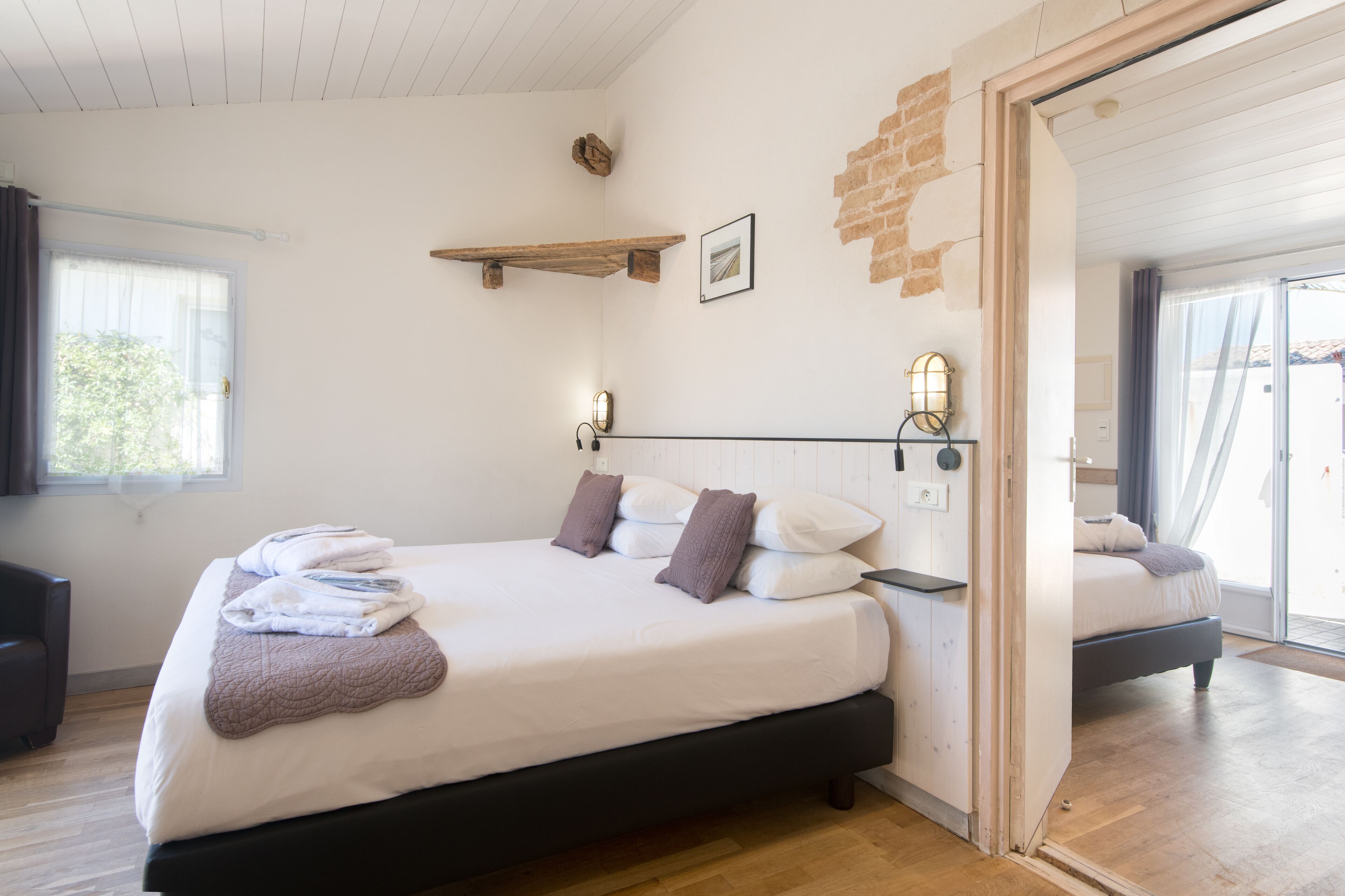 A bedroom with a bed, a wooden headboard, a bedside table, a wall-mounted shelf, and a window with curtains.