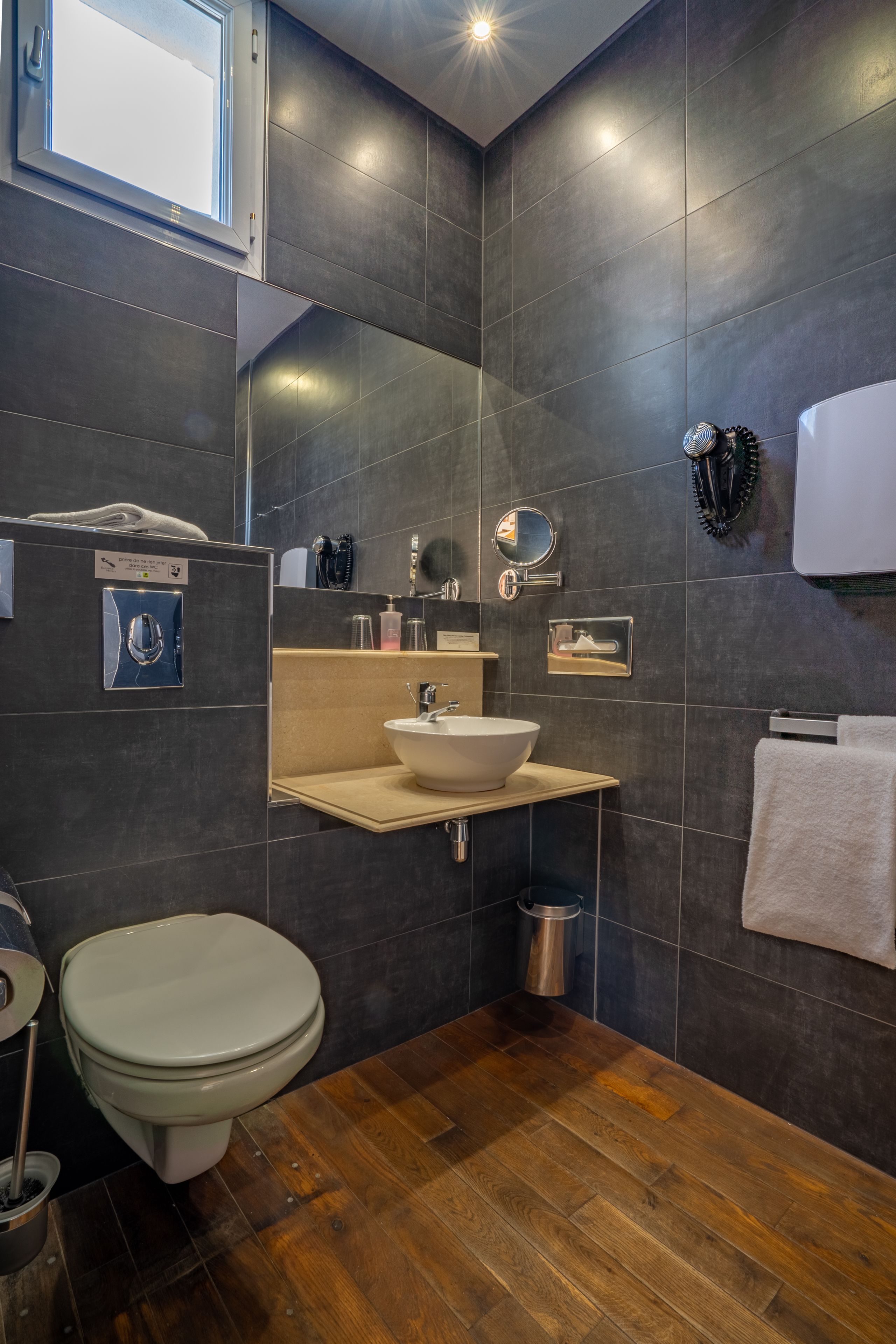A modern bathroom with dark tiles, a wooden floor, a white sink, a toilet, and a mirror.