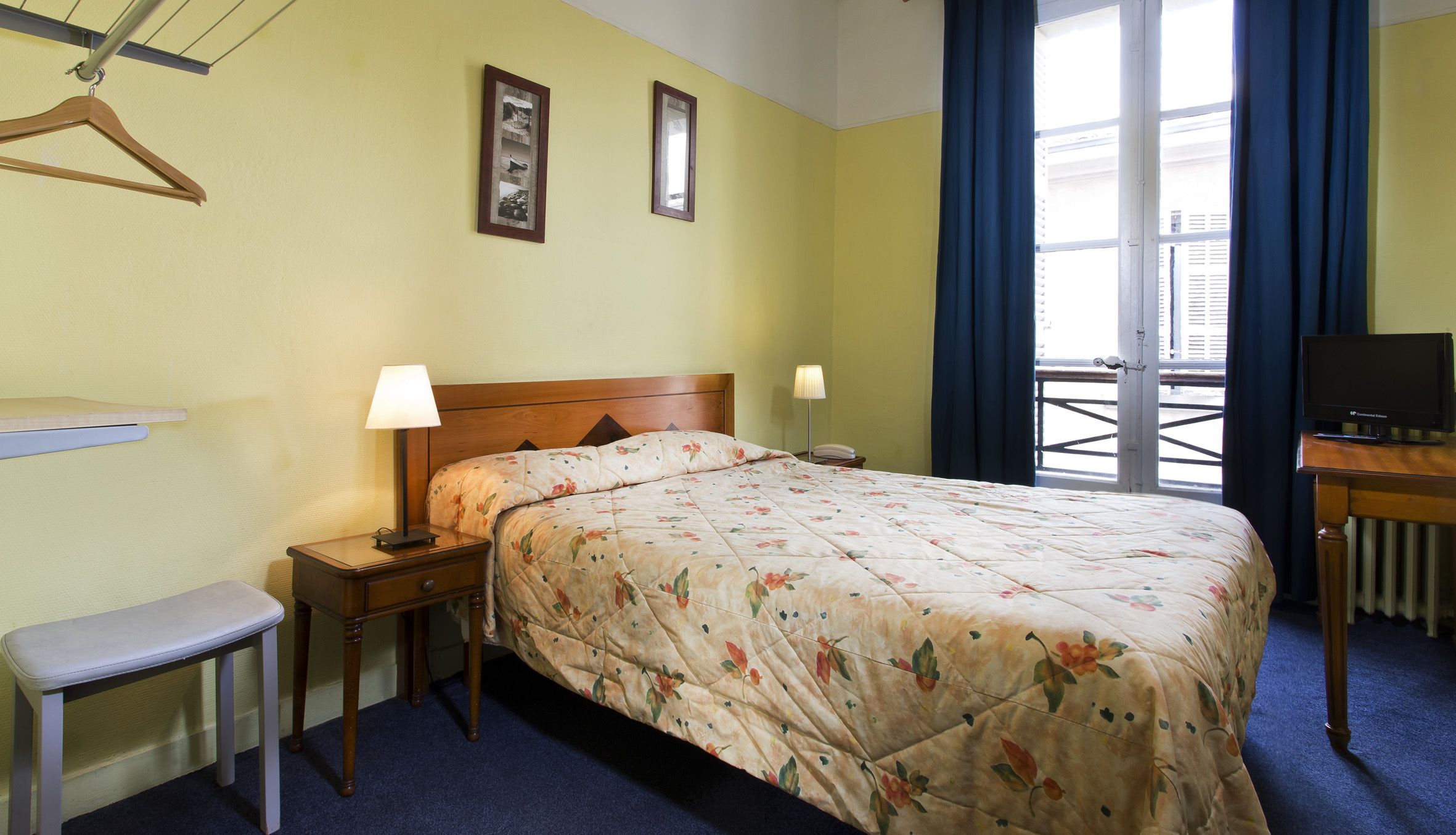 Habitación de hotel con cama, mesitas de noche, un banco, televisor y ventana con cortinas.