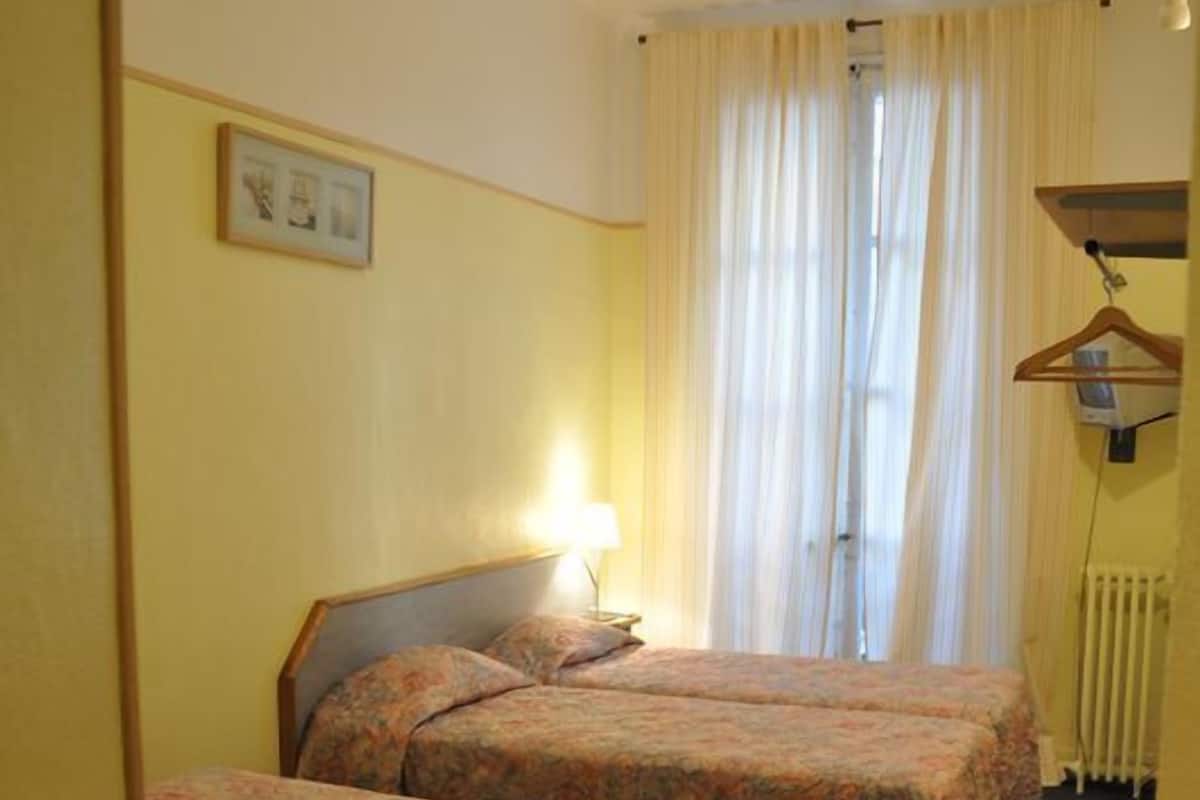 A hotel room with two single beds, a small desk, a chair, a lamp, and a radiator.