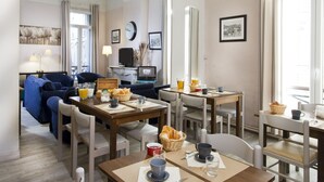 A modern hotel room with a dining area, a sofa, a TV, and a dining table set with breakfast items. - Hotel Sylvabelle (Marseille)