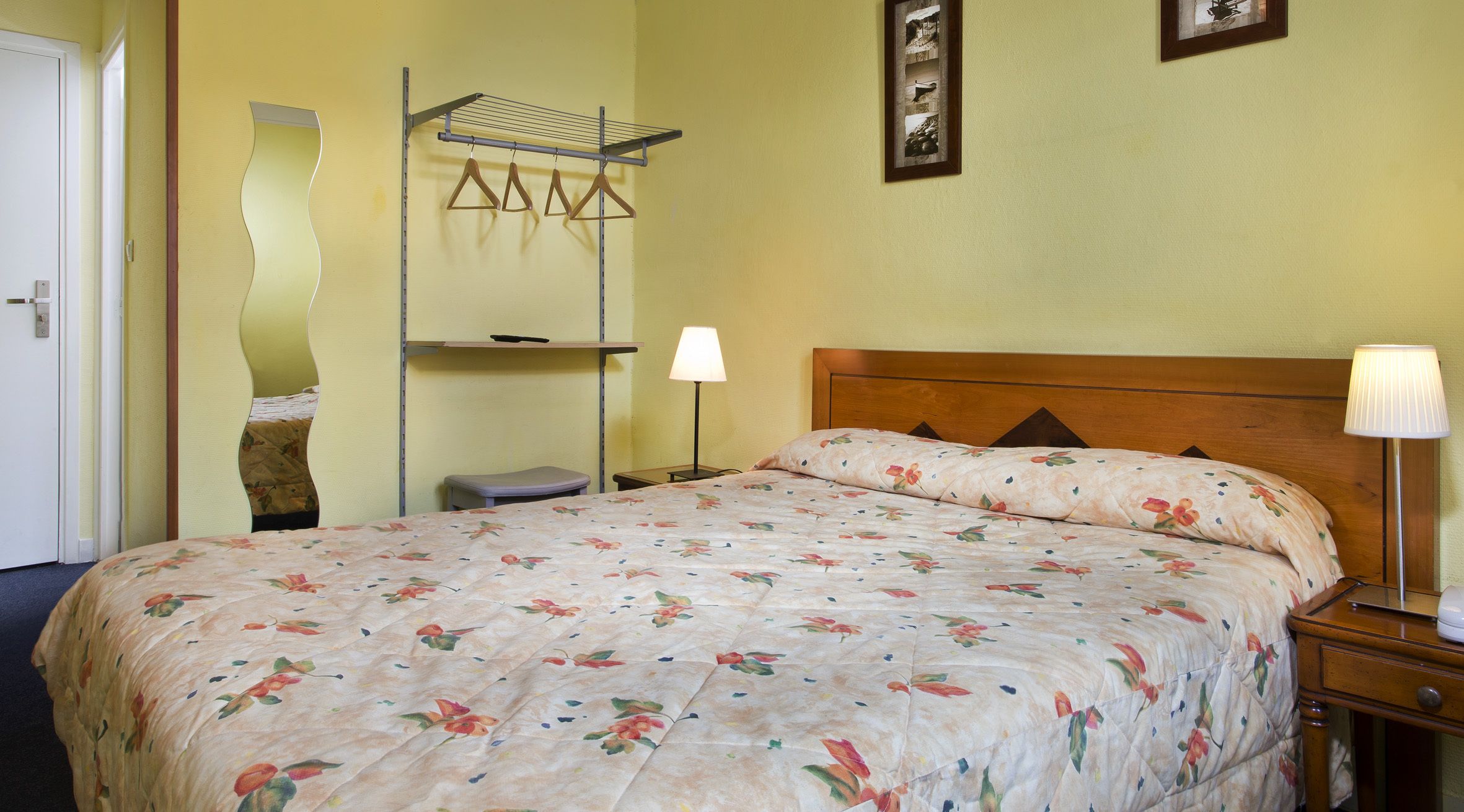 Una cama bien hecha con una colcha estampada con flores, una lámpara de noche y un tendedero de pared.