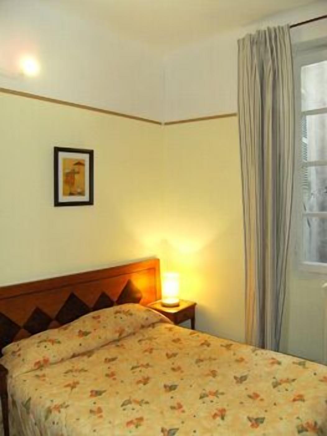 A hotel room with a bed, a nightstand with a lamp, a framed picture on the wall, and a window with curtains.