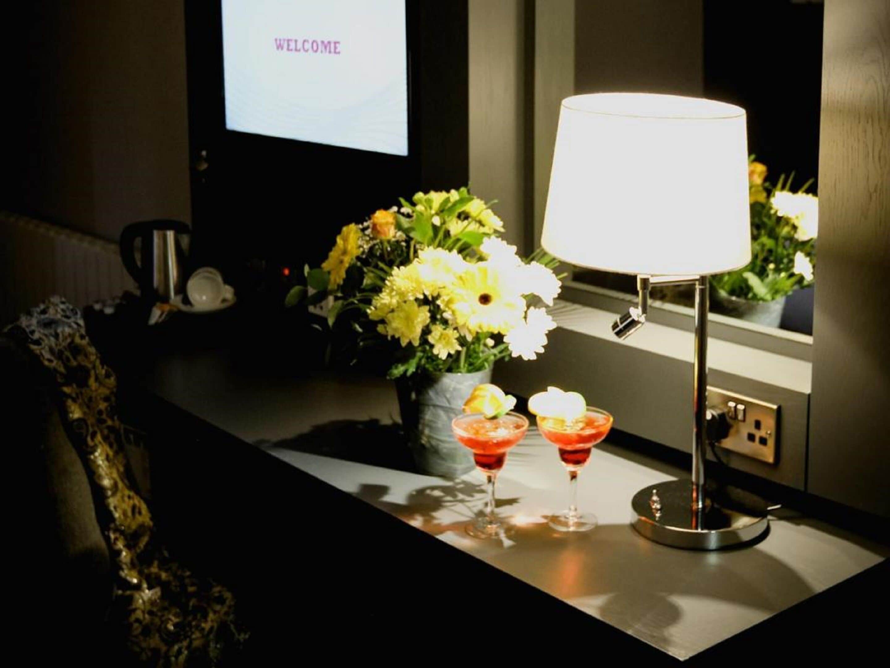 Un'area reception con schermo di benvenuto, un tavolo con lampada, fiori e due bicchieri.