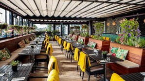 Un restaurante al aire libre con mesas y sillas de madera, toallas amarillas y plantas colgantes.