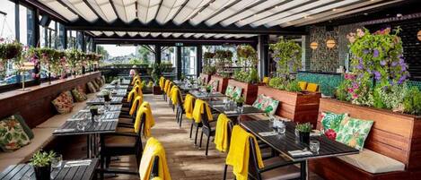 Un restaurante al aire libre con mesas y sillas de madera, toallas amarillas y plantas colgantes.
