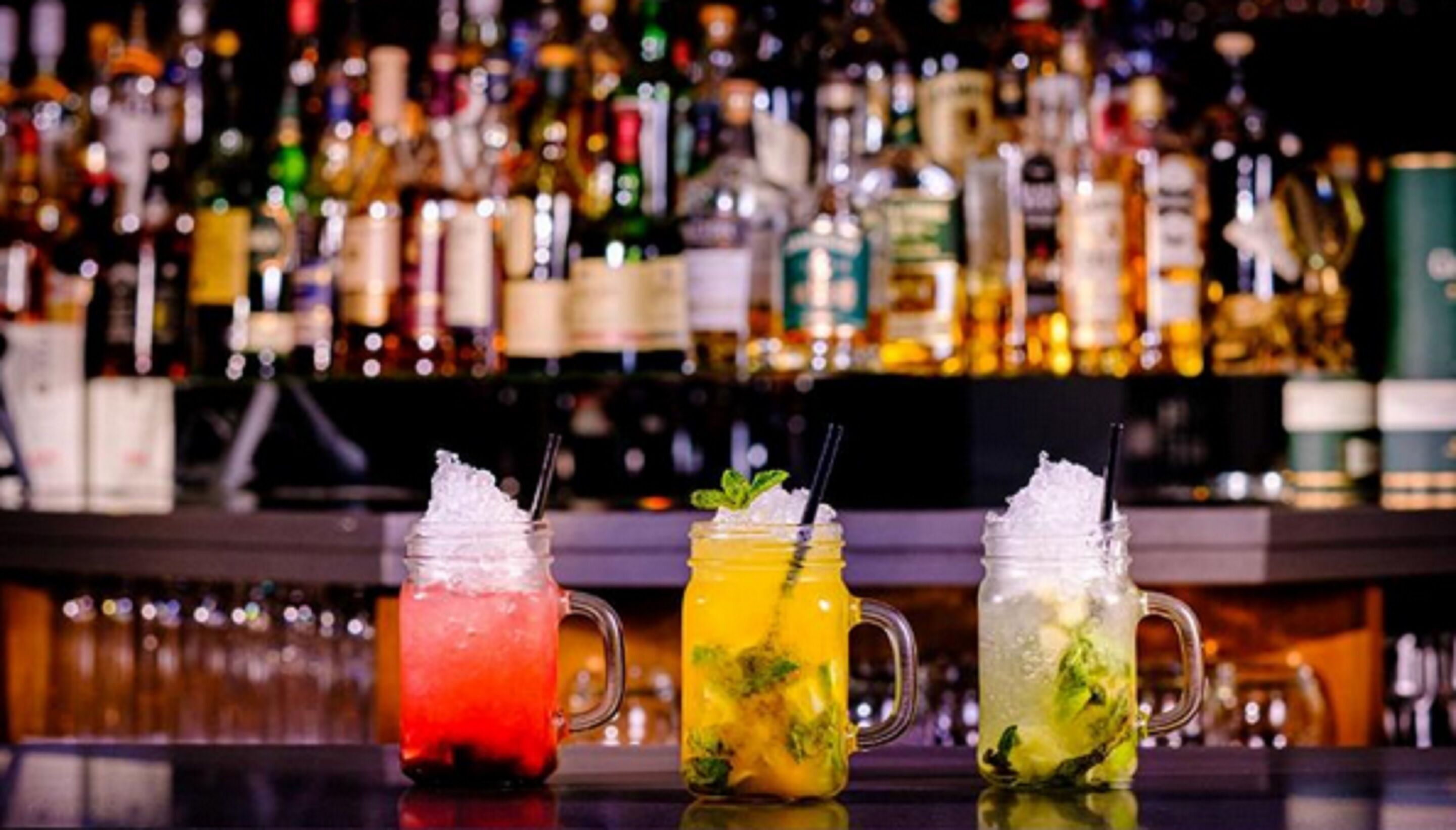 Drei bunte Cocktails auf einer Bartheke vor einer Vielzahl von Flaschen.