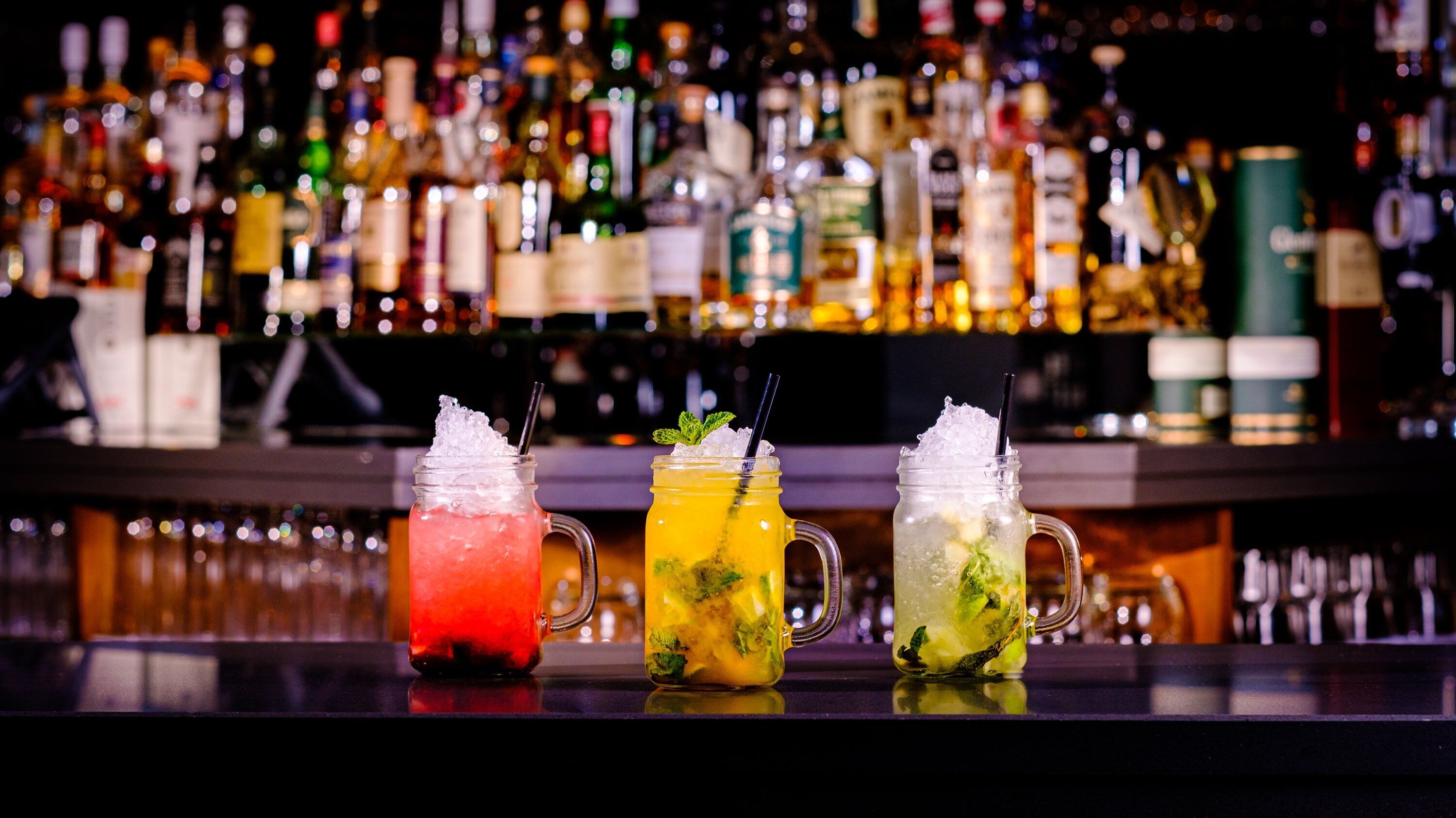 Three colorful cocktails on a bar counter with a blurred background of various bottles.