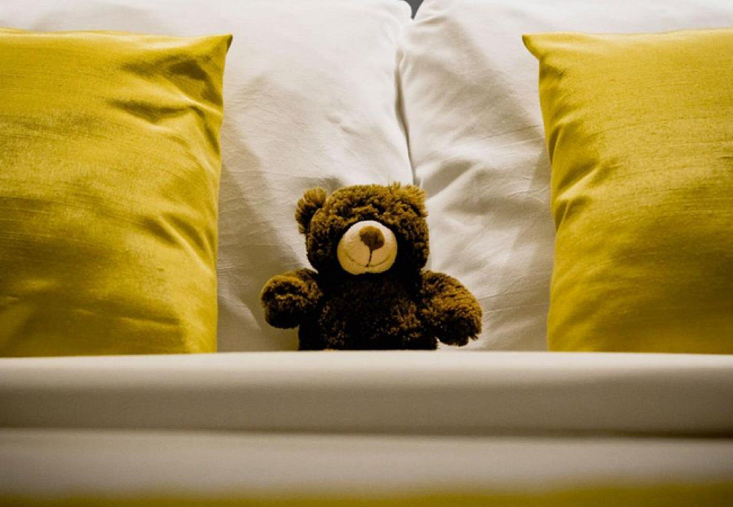 A bed with a white mattress and two yellow pillows, a brown teddy bear in the center.