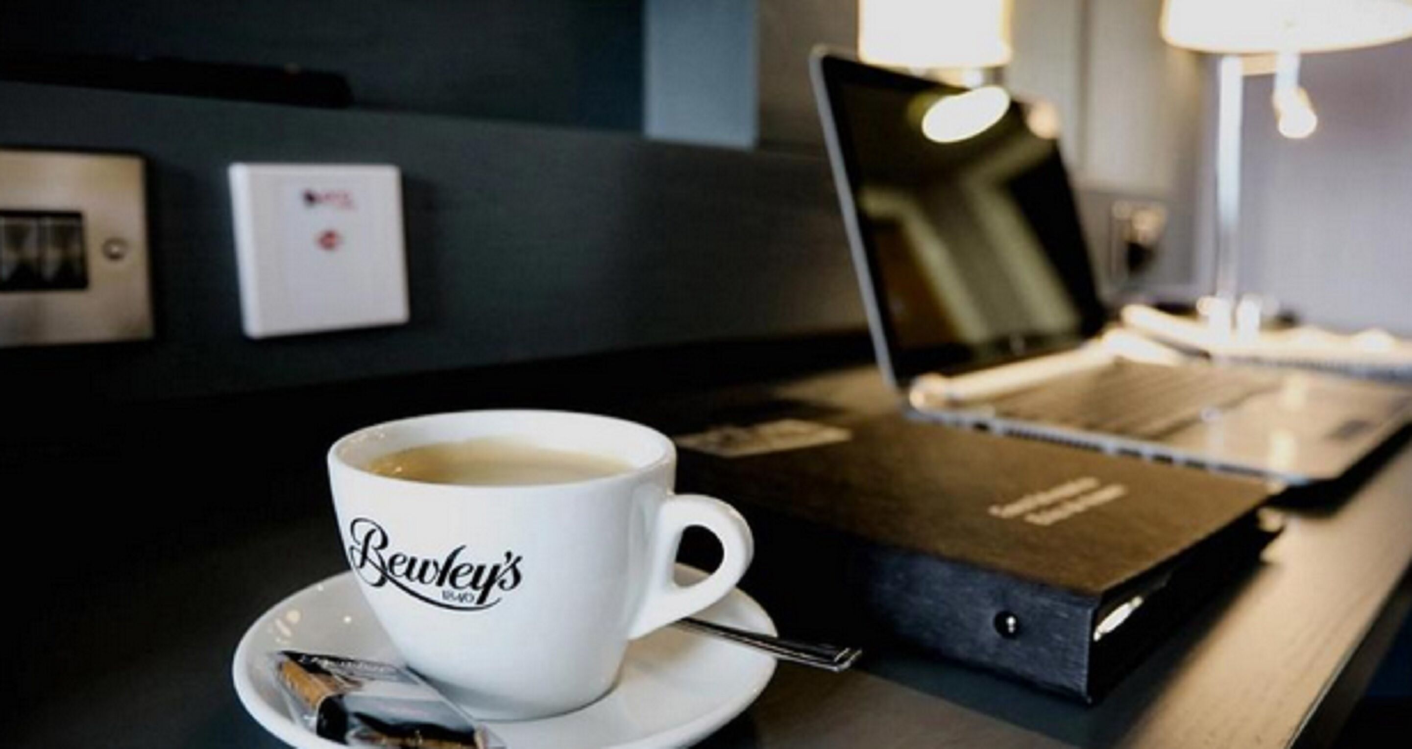 A cup of coffee on a saucer with a spoon, a laptop, and a box on a table.