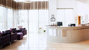 Un moderno vestíbulo de hotel con un mostrador de recepción curvo, zona de estar con sofás morados, ventanas amplias con cortinas traslúcidas y una decoración minimalista.