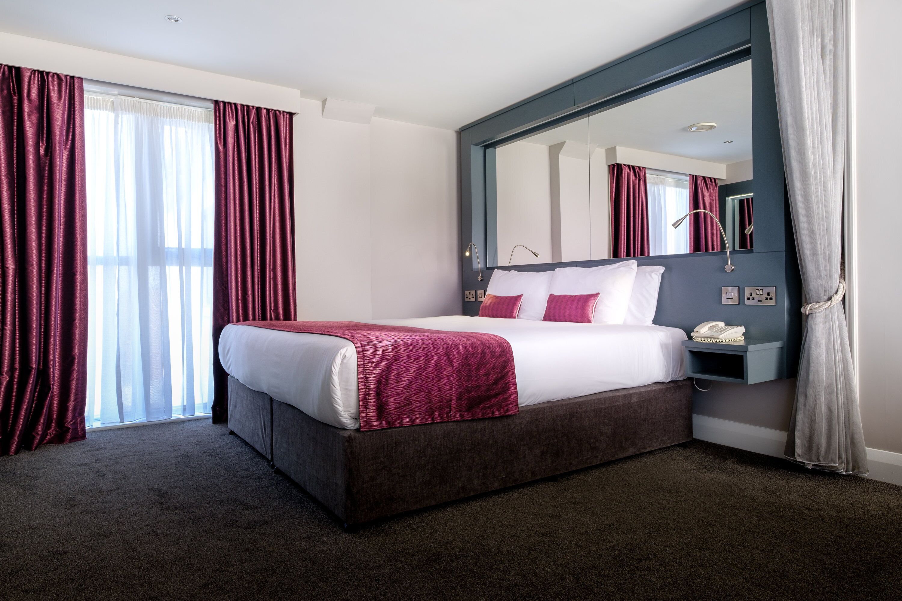 A hotel room with a large bed, a bedside table with a phone, and a mirror above the bed.