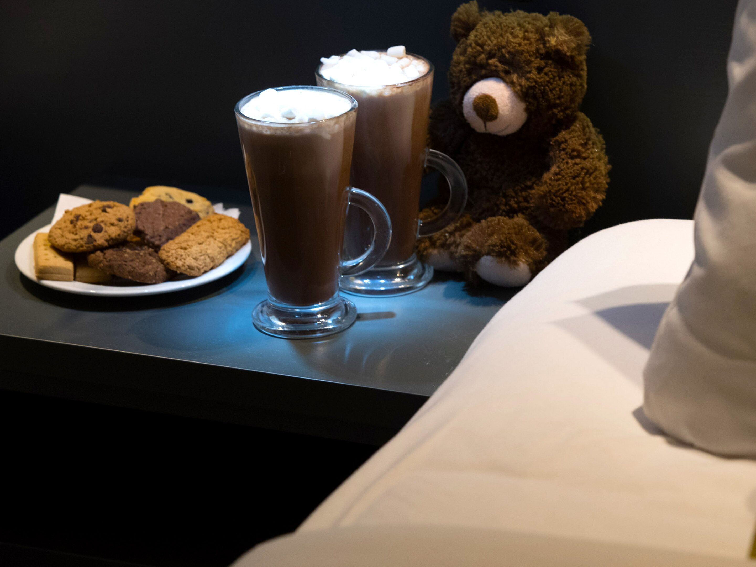 A table with two glasses of hot chocolate topped with marshmallows, a plate of cookies, and a teddy bear.