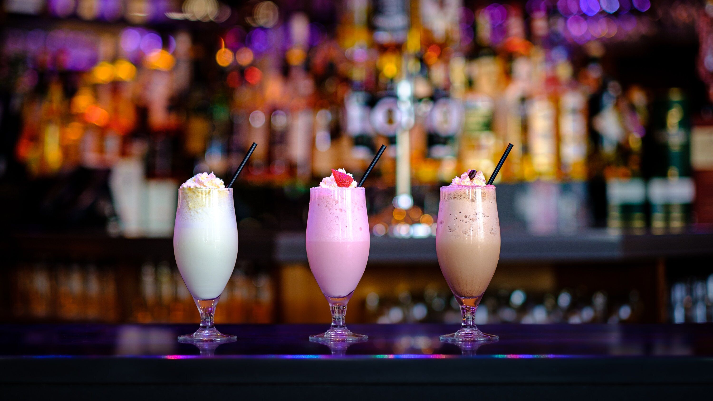 Trois verres posés sur le comptoir d’un bar, avec un arrière-plan flou composé de bouteilles.