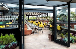 Un área de comedor al aire libre con mesas y sillas, algunas cubiertas con telas amarillas, y una variedad de plantas alrededor.