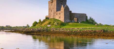 A historic stone castle with a large tower, situated on a grassy hill overlooking a calm body of water.
