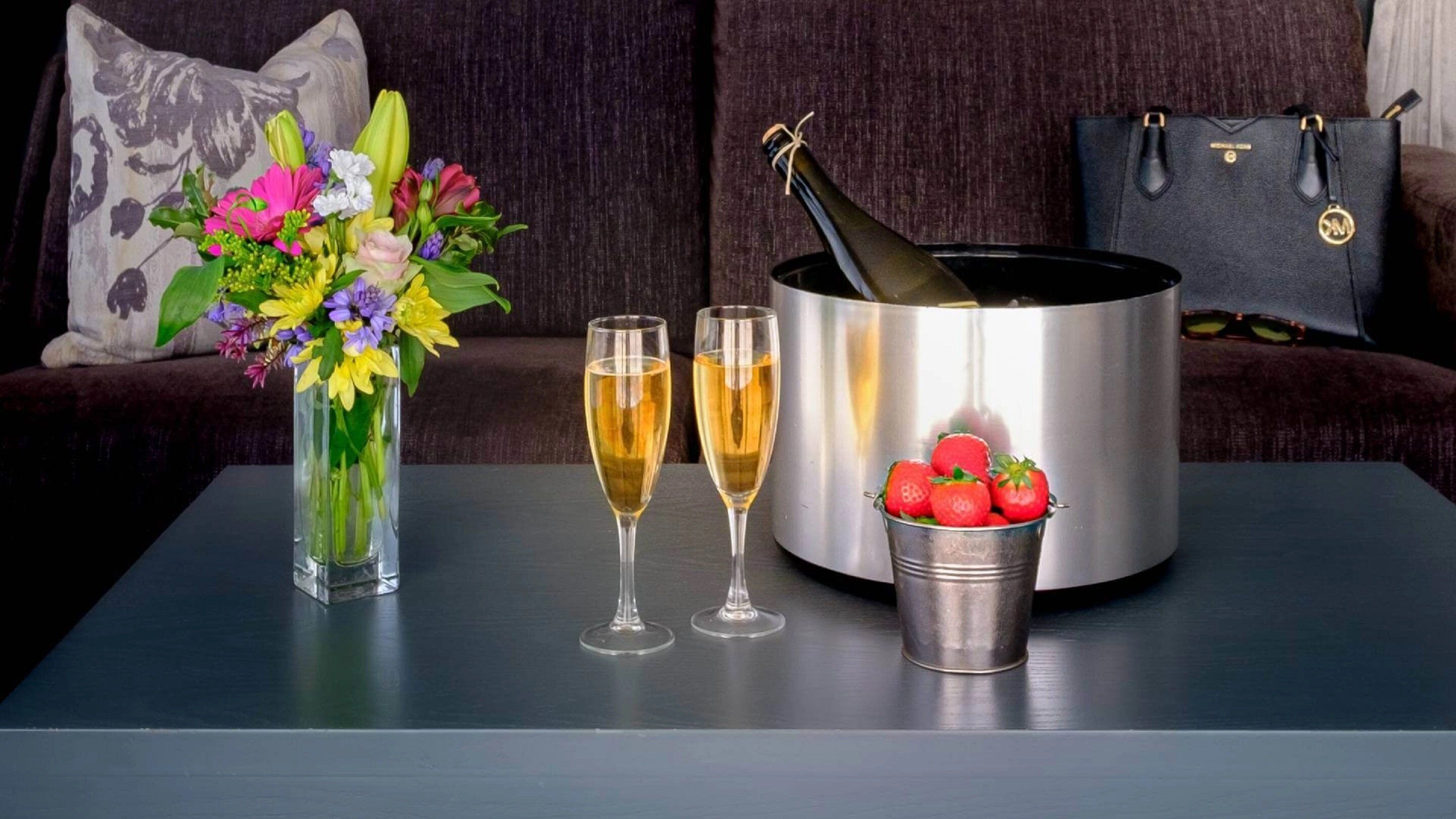 Una mesa con una botella de champán, dos copas de champán, un cubo de fresas y un jarrón de flores.