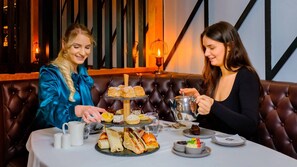 Dos personas sentadas a una mesa con diversos pasteles y sándwiches, una tetera y una taza.