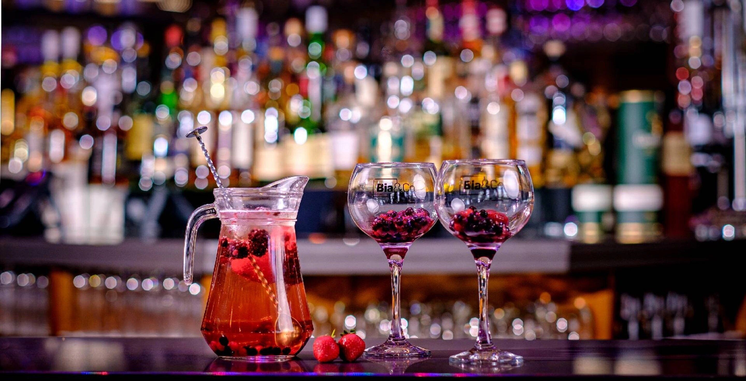 Un bar con un jarro y dos vasos llenos de una bebida roja, frutos rojos y una pajita.