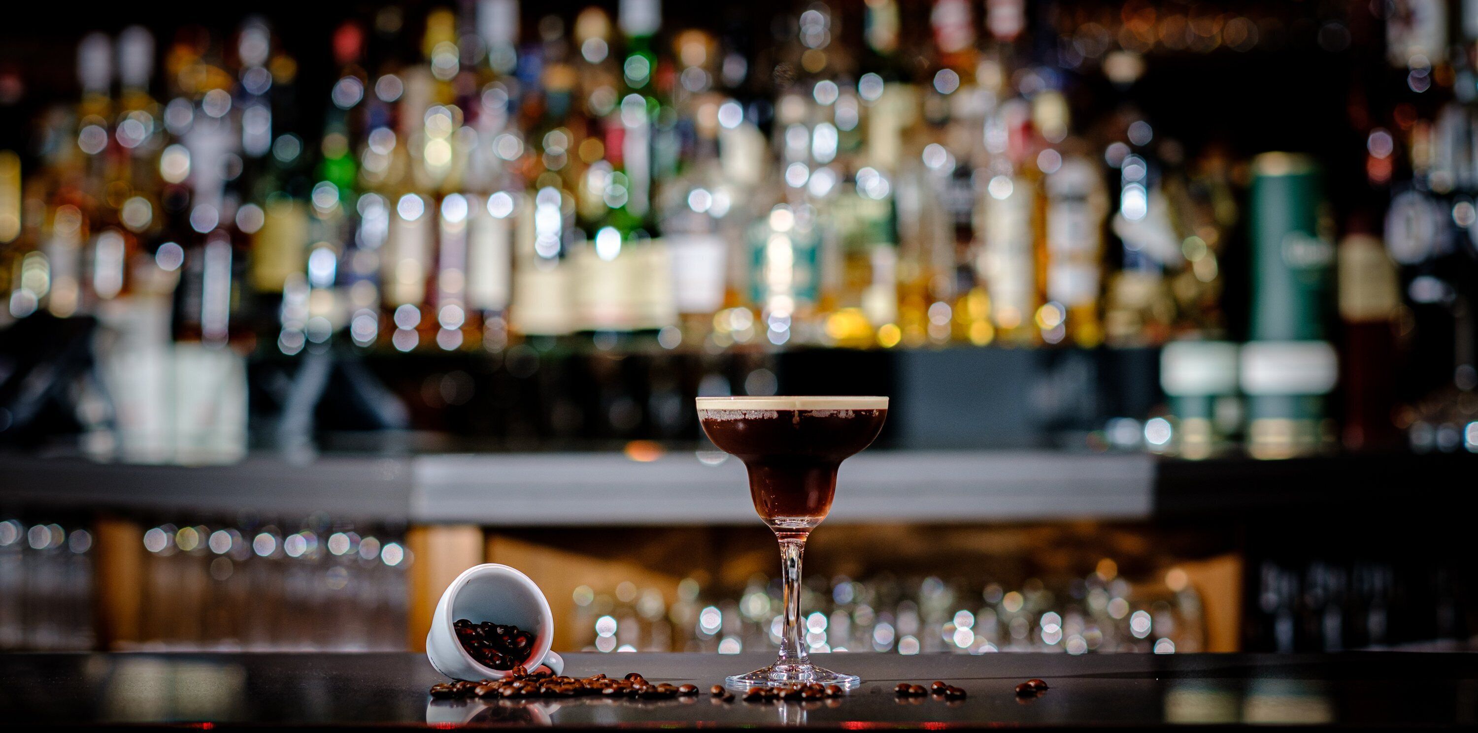 A bar setting with a cocktail glass and a coffee cup on the counter, surrounded by various bottles in the background.