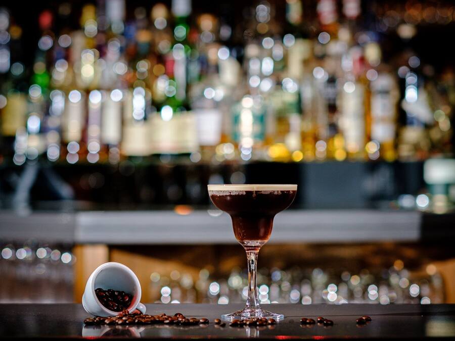 A bar setting with a cocktail glass and a coffee cup on the counter, surrounded by various bottles in the background.
