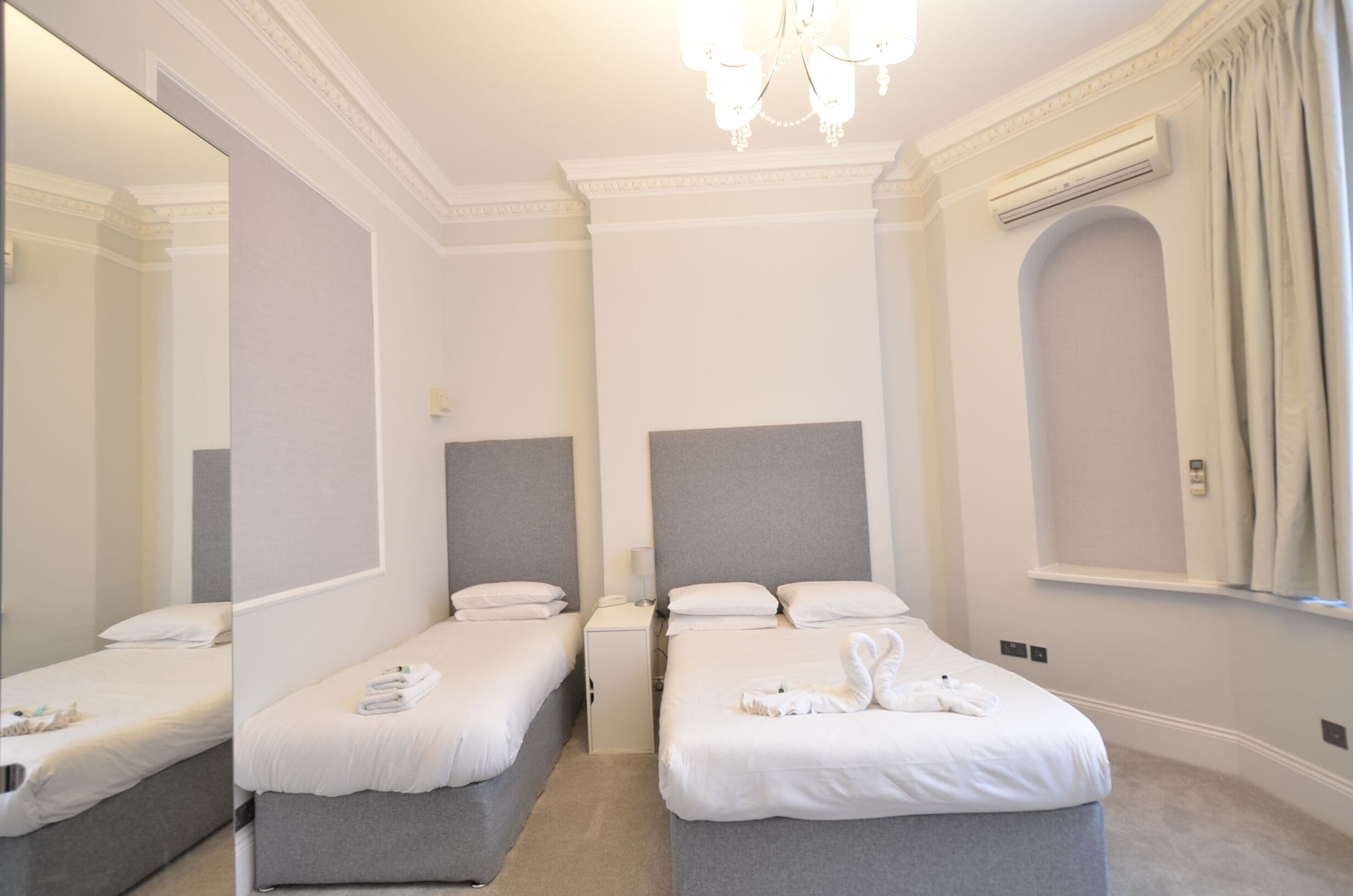 A hotel room with two beds, a large mirror, a chandelier, and a window with curtains.